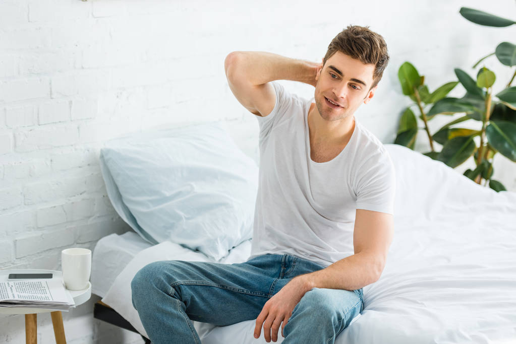 przystojny mężczyzna w t-shirt i dżinsy, siedząc na łóżku w sypialni - Zdjęcie, obraz
