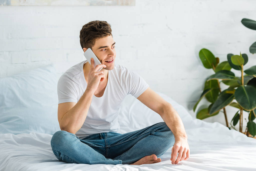 Mann in T-Shirt und Jeans sitzt auf Bett und telefoniert im Schlafzimmer mit Smartphone - Foto, Bild