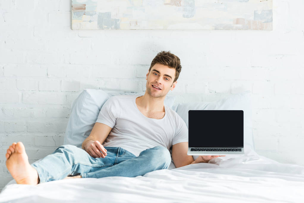 schöner Mann in weißem T-Shirt und Jeans sitzt auf dem Bett und hält Laptop mit leerem Bildschirm im Schlafzimmer - Foto, Bild