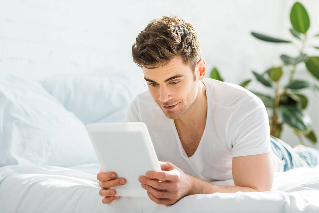 knappe man in de witte t-shirt liggend op bed met digitale tablet in slaapkamer - Foto, afbeelding