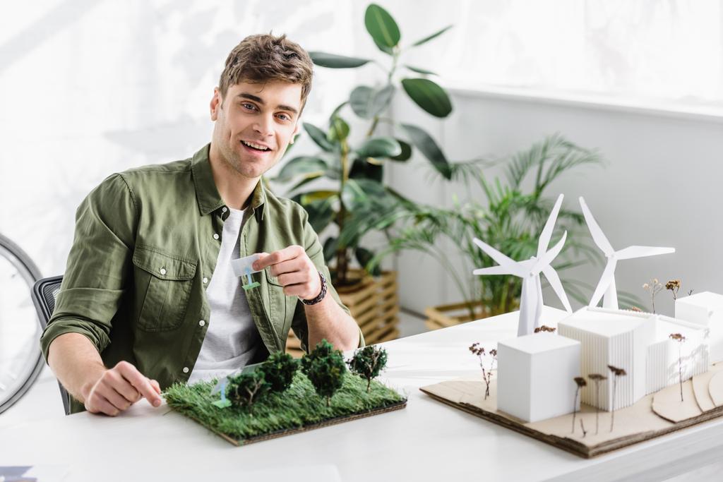 schöner Architekt lächelt und hält Solarmodellen über Gras auf dem Tisch im Büro - Foto, Bild