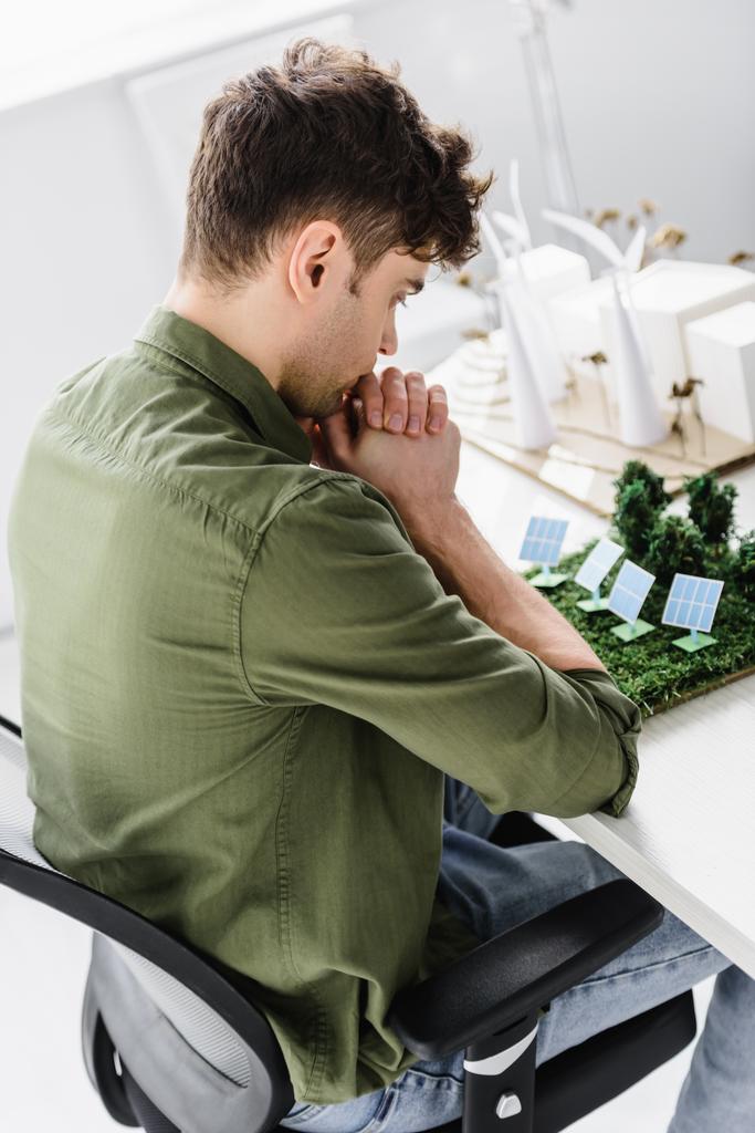 handsome architect sitting on chair at table with trees, windmills, buildings and solar panels models in office - Photo, Image
