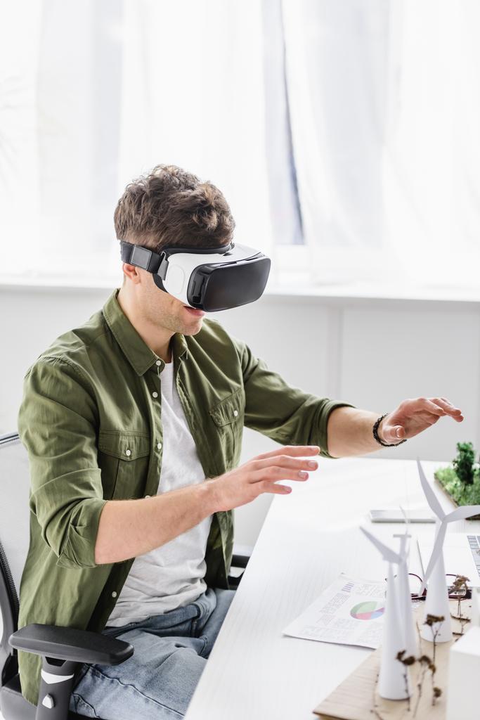 архітектор у гарнітурі віртуальної реальності, що сидить за столом з вітряками та деревами в офісі
 - Фото, зображення