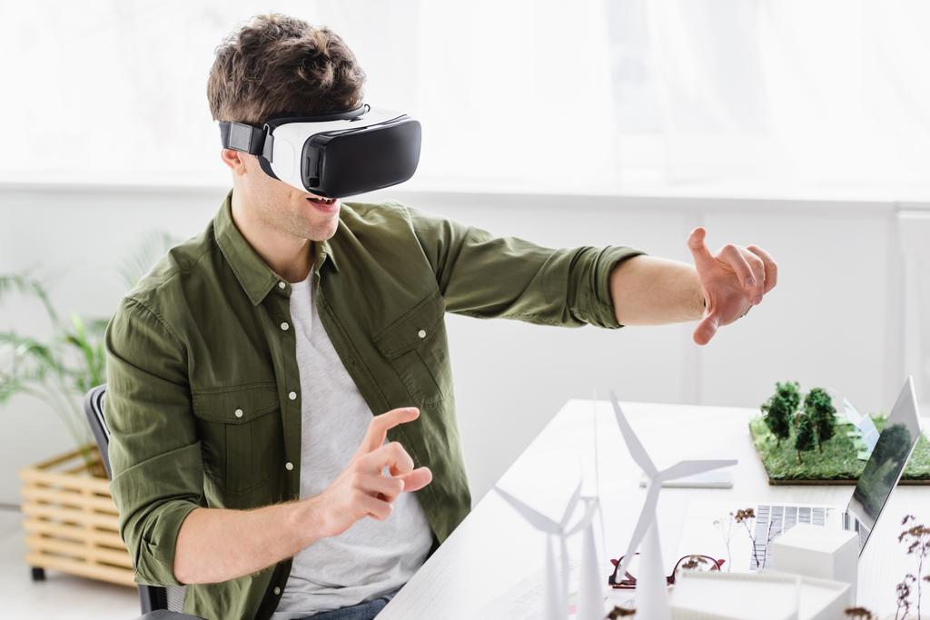architect in virtual reality headset sitting at table with laptop, windmills and trees models in office - Photo, Image