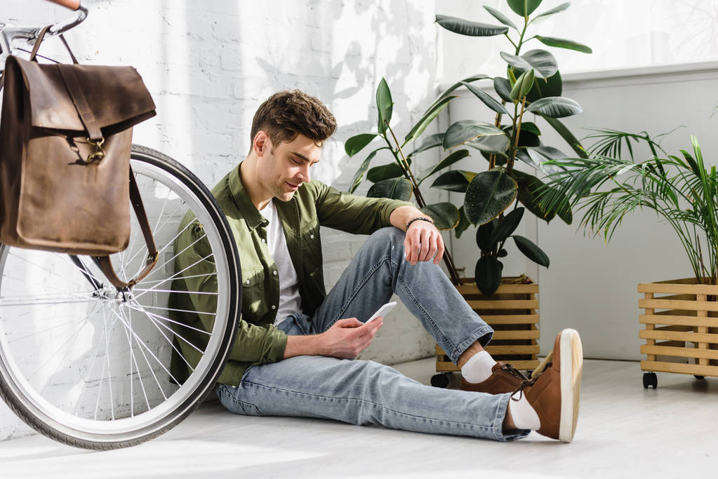 man in groene shirt en spijkerbroek bedrijf smartphone, zitten in de buurt van bakstenen muur-, fiets- en planten in kantoor - Foto, afbeelding