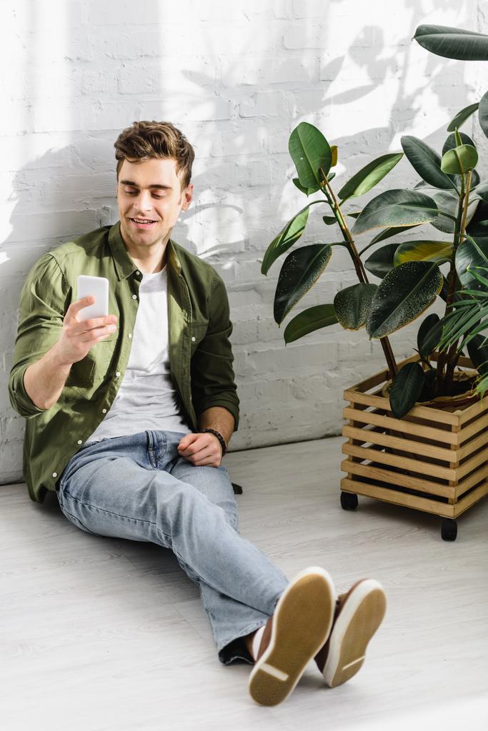 man in groene shirt en spijkerbroek bedrijf smartphone, zitten in de buurt van bakstenen muur en planten in kantoor - Foto, afbeelding