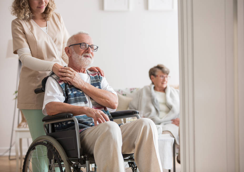 Старший мужчина в инвалидном кресле при содействии молодой медсестры
 - Фото, изображение