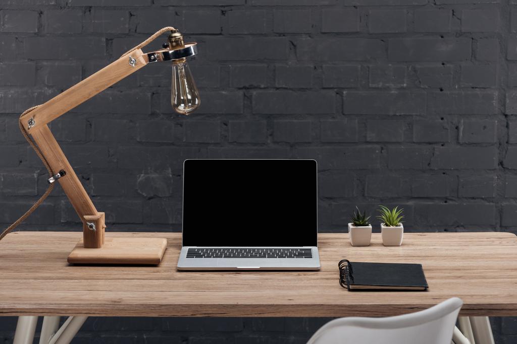 moderne werkplaats met laptop met leeg scherm, planten, lamp en notitieblok op houten tafel  - Foto, afbeelding