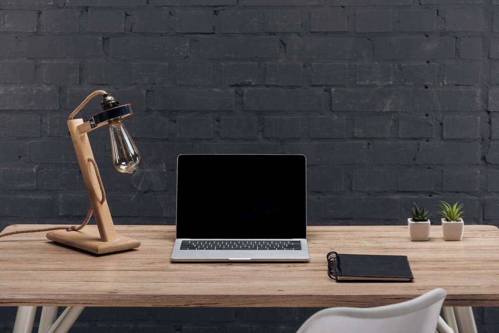 moderner Arbeitsplatz mit Laptop mit leerem Bildschirm, Dekor, Lampe und Notizbuch auf Holztisch neben schwarzer Ziegelwand - Foto, Bild