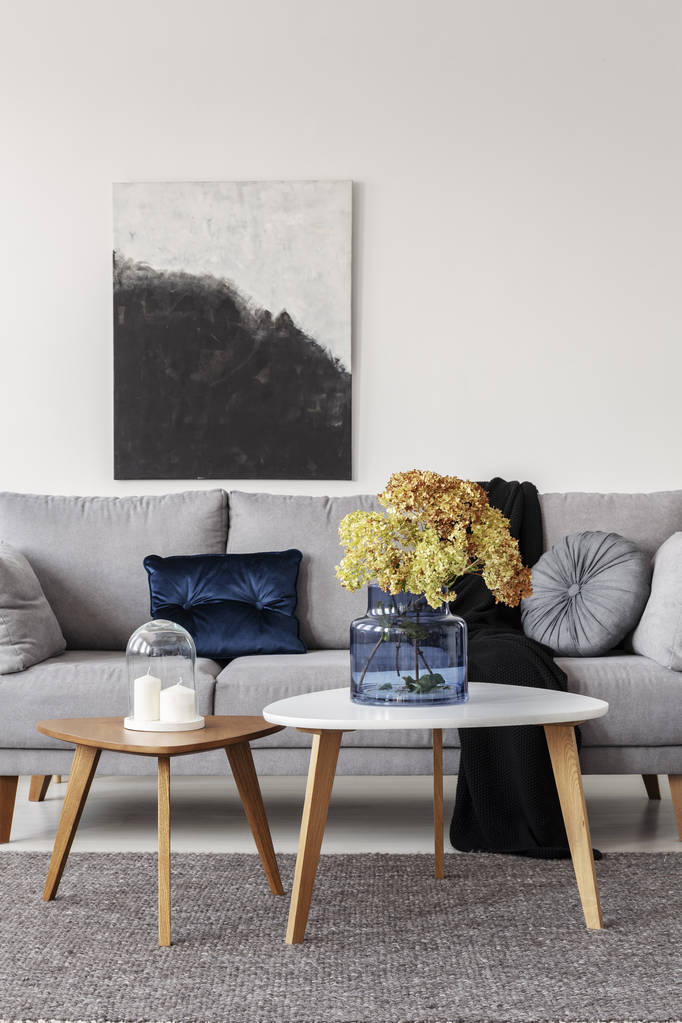 Blumen in blauer Glasvase und zwei weiße Kerzen auf Holztischen im grau-eleganten Wohnzimmer mit gemütlichem Sofa - Foto, Bild