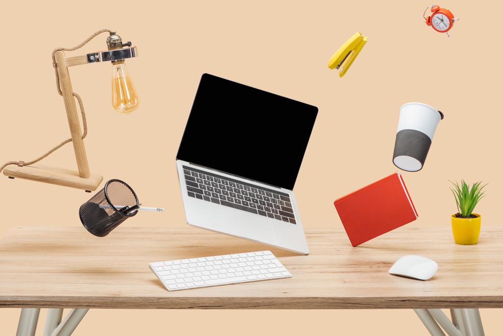 Laptop mit leerem Bildschirm und Schreibwaren schweben in der Luft über Holztisch isoliert auf beige - Foto, Bild