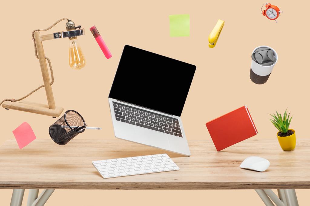 laptop met leeg scherm, lamp, kleverige nota's en briefpapier zwevende in de lucht boven houten bureau geïsoleerd op beige - Foto, afbeelding