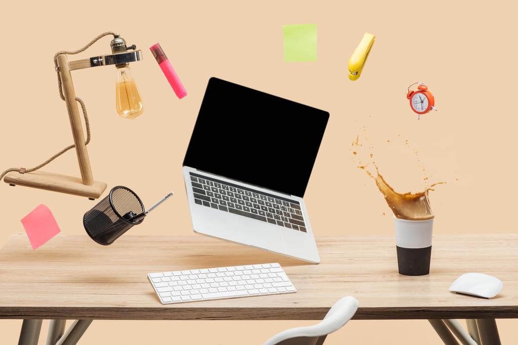 laptop with blank screen, lamp, empty sticky notes and stationery levitating in air above workplace with thermomug with coffee splash  on table isolated on beige - Photo, Image