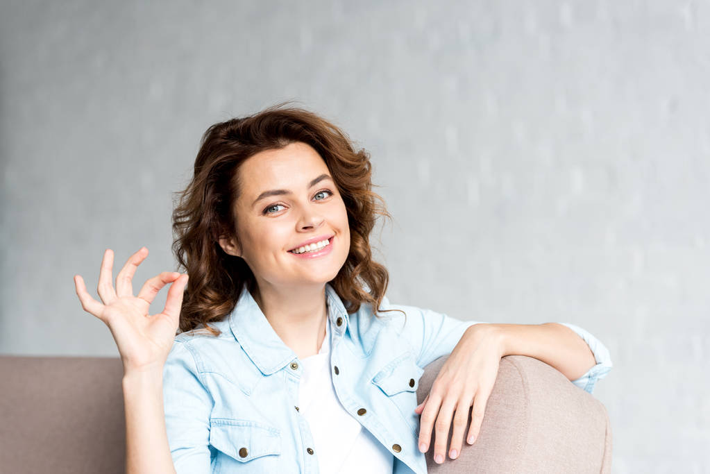 Mulher encaracolado feliz na camisa sentado no sofá e mostrando sinal ok em cinza
  - Foto, Imagem