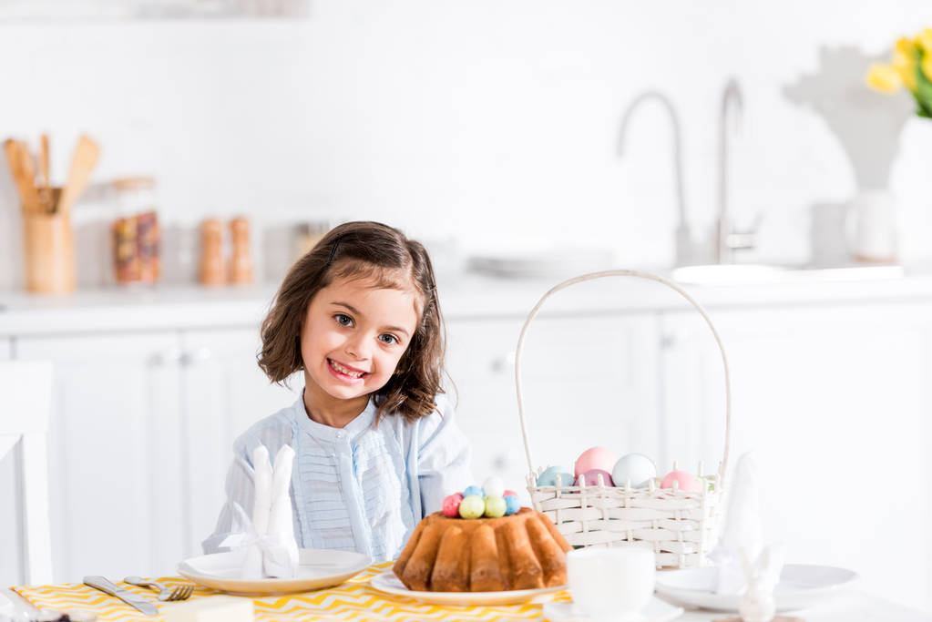 Niño adorable sentado a la mesa con pastel de Pascua y huevos pintados en la cocina
 - Foto, Imagen