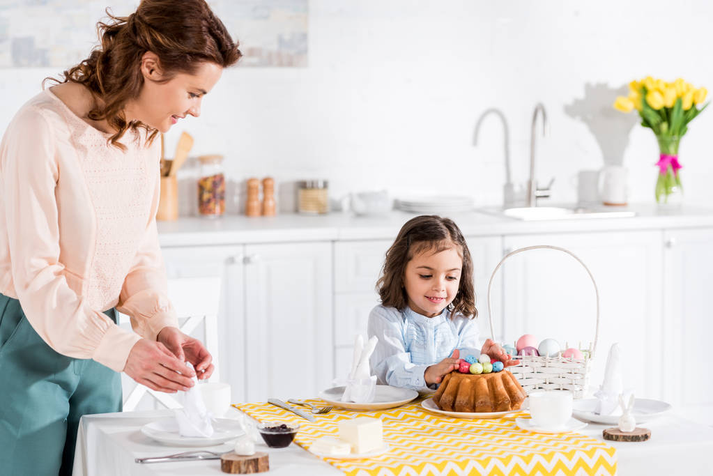 Criança sorridente olhando para bolo de Páscoa enquanto a mãe dobra guardanapos
 - Foto, Imagem