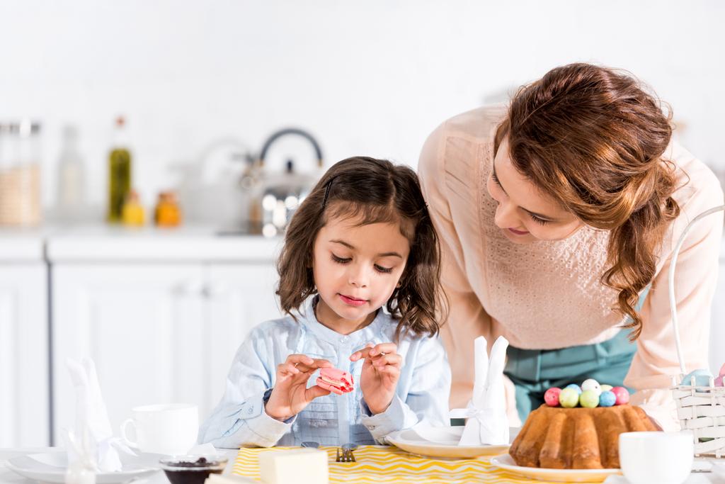 Wesoły kobieta patrząc na córkę jedzenie w kuchni makaronik - Zdjęcie, obraz
