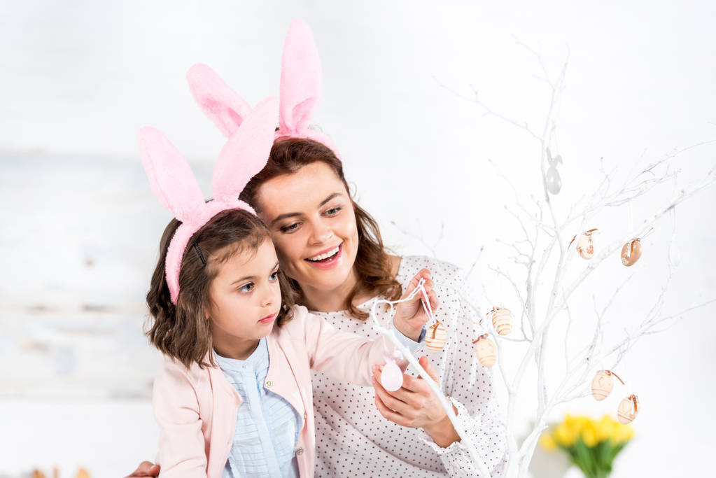 Blissful mother and daughter in bunny ears decorating easter tree - Photo, Image
