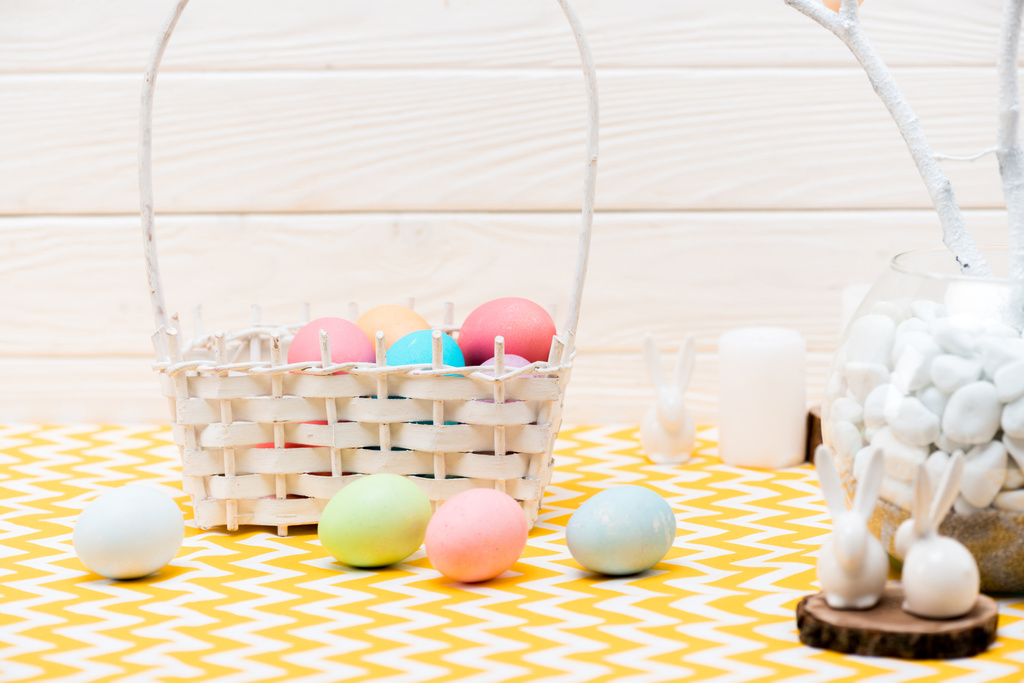 Плетеная корзина с окрашенными пасхальными яйцами и керамическими кроликами на столе
 - Фото, изображение