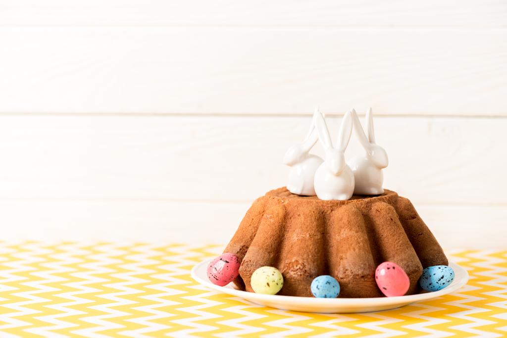Пасхальный торт с окрашенными перепелиными яйцами и керамическими кроликами на столе
 - Фото, изображение