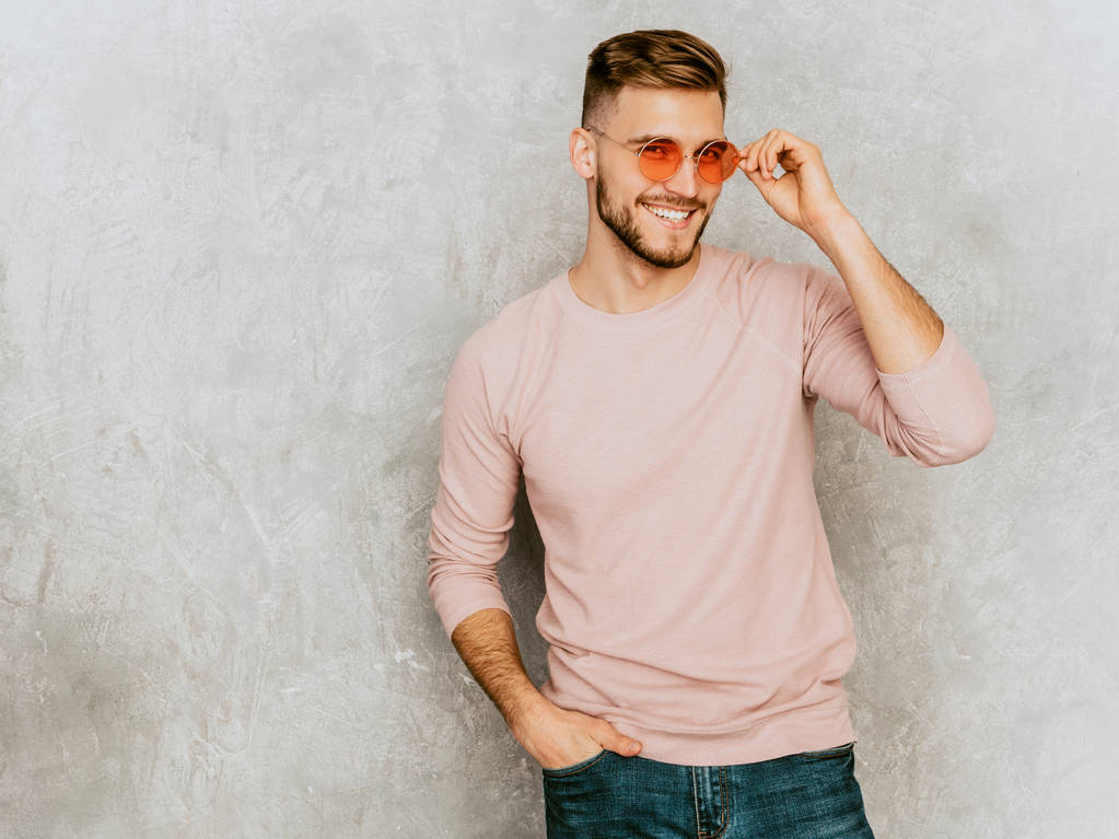 Porträt eines hübschen lächelnden Hipster-Geschäftsmann-Models in lässiger sommerlicher rosa Kleidung. Modebewusster Mann posiert mit runder Sonnenbrille vor grauer Wand - Foto, Bild