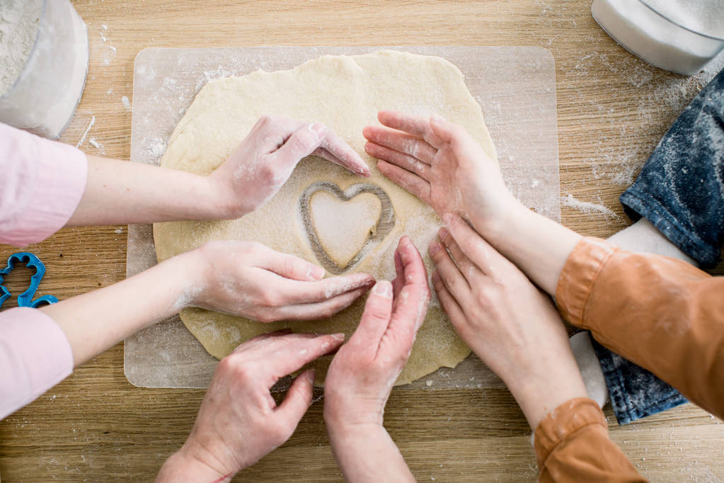 cooking and home concept - close up of female hands making cookies from fresh dough at home. Hands of three women hold cookie in form of heart - Photo, Image
