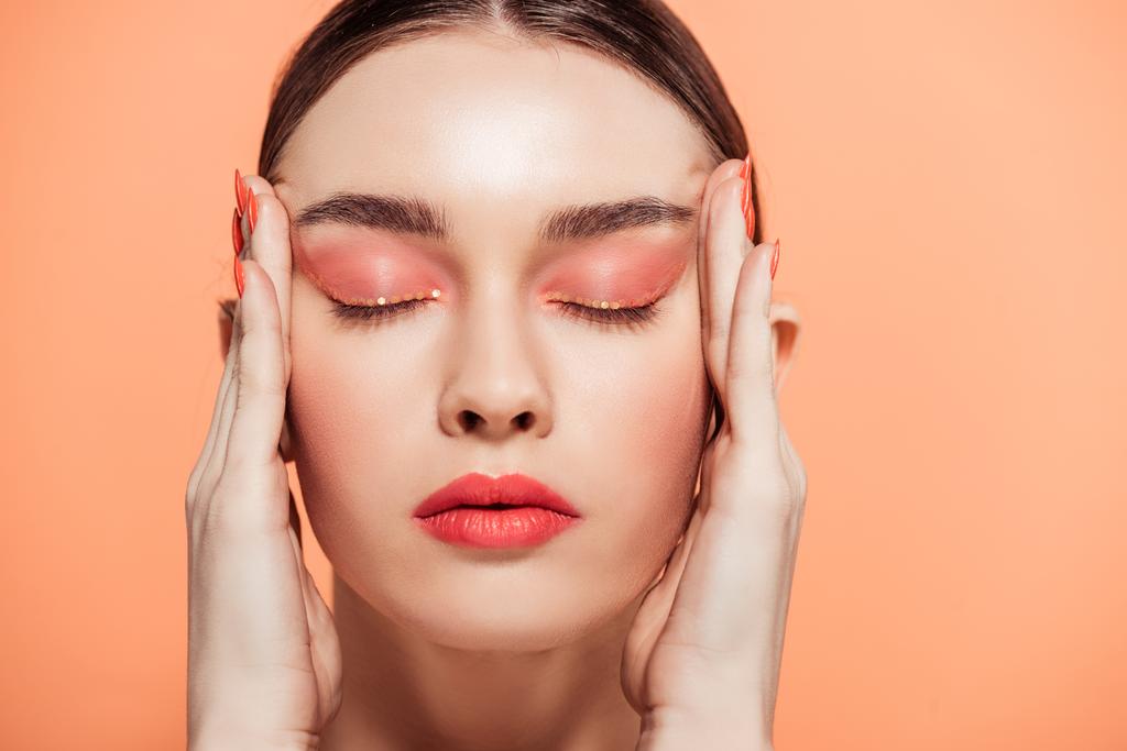 beautiful trendy young woman with glitter makeup and eyes closed touching face isolated on coral - Photo, Image