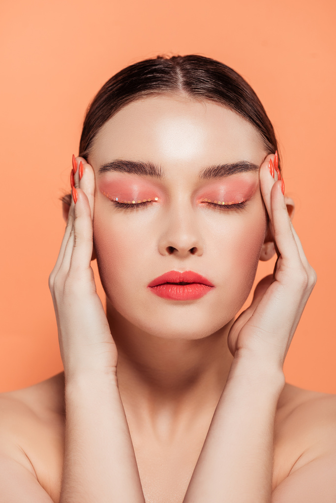 mooie trendy jonge vrouw met glitter make-up en ogen gesloten aanraken van gezicht geïsoleerd op koraal - Foto, afbeelding