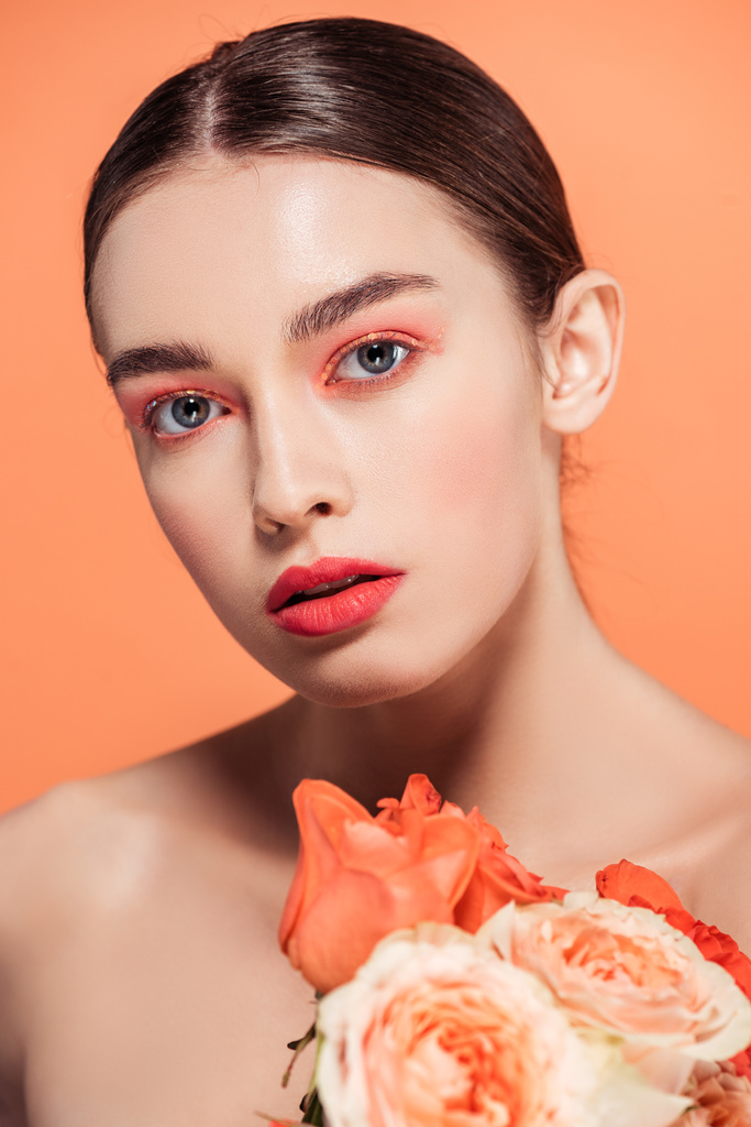 hermosa chica con estilo mirando a la cámara y posando con flores de rosas aisladas en coral
 - Foto, Imagen