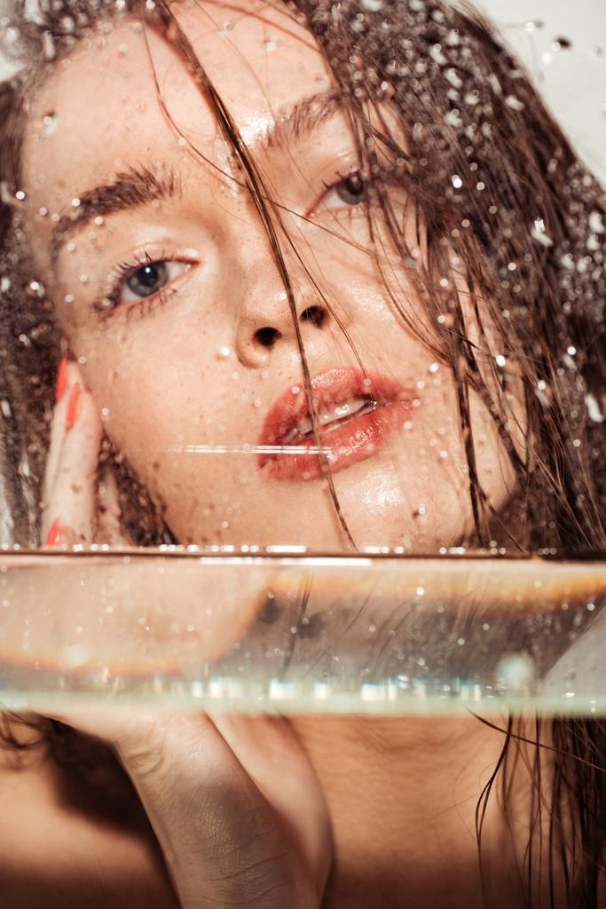 όμορφη νεαρή γυναίκα με κοράλλι χείλη κοιτάζοντας την κάμερα μέσα από γυαλί με σταγόνες νερού - Φωτογραφία, εικόνα