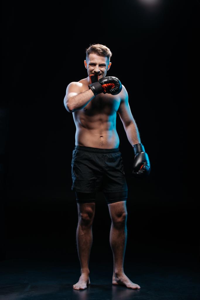 muscular barefoot sporty boxer taking off  boxing gloves with mouth on black - Photo, Image