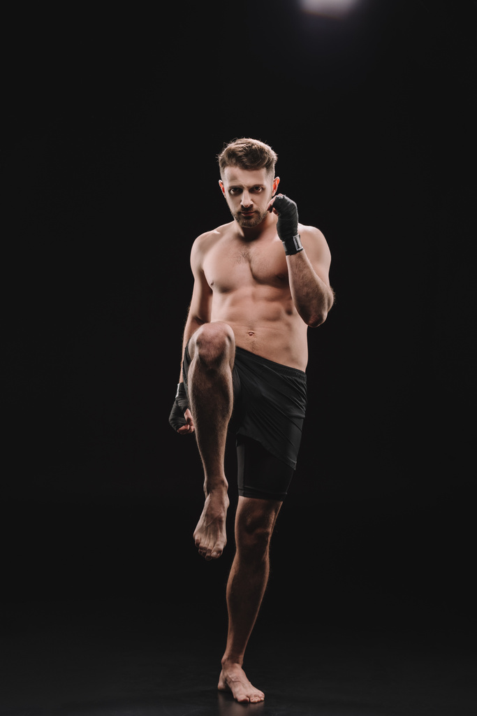 vista frontale di muscoloso a piedi nudi mma combattente in bende facendo calcio con il ginocchio sul nero
 - Foto, immagini