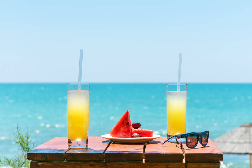 Dois copos de suco de frutas tropicais frescas na praia, e um prato com uvas e melancia na mesa, contra o mar. Conceito de férias e férias
 - Foto, Imagem