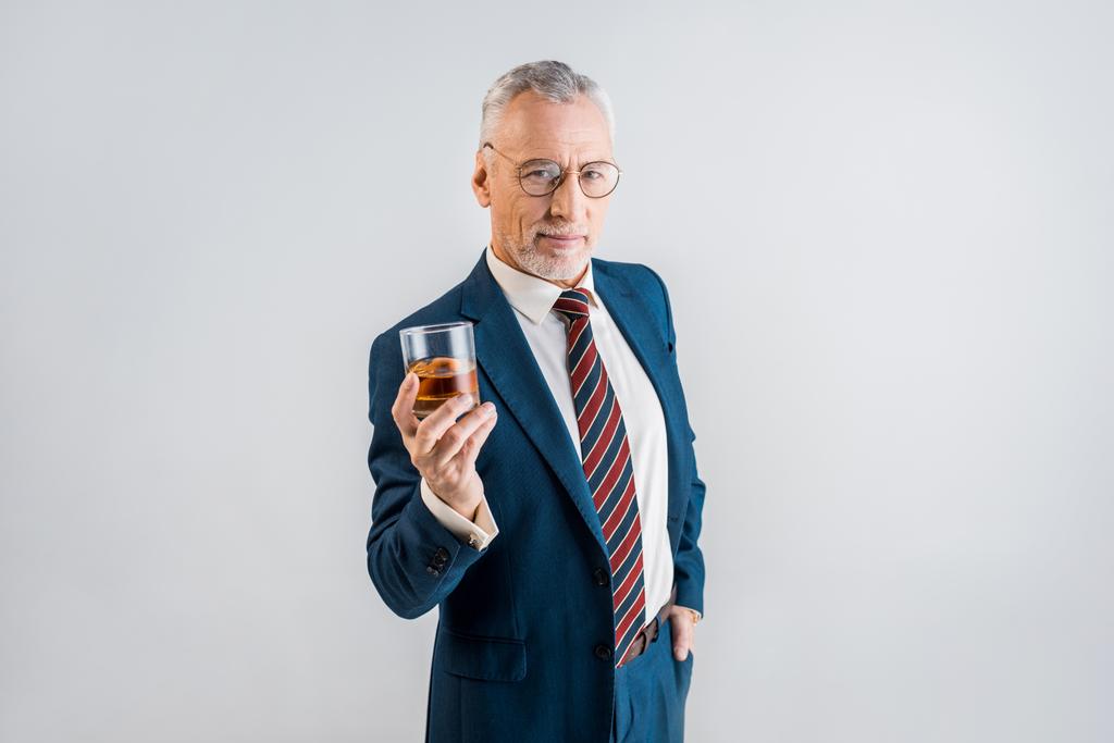 Veselý starší podnikatel drží sklenici whisky stoje s rukou v kapse izolované Grey  - Fotografie, Obrázek