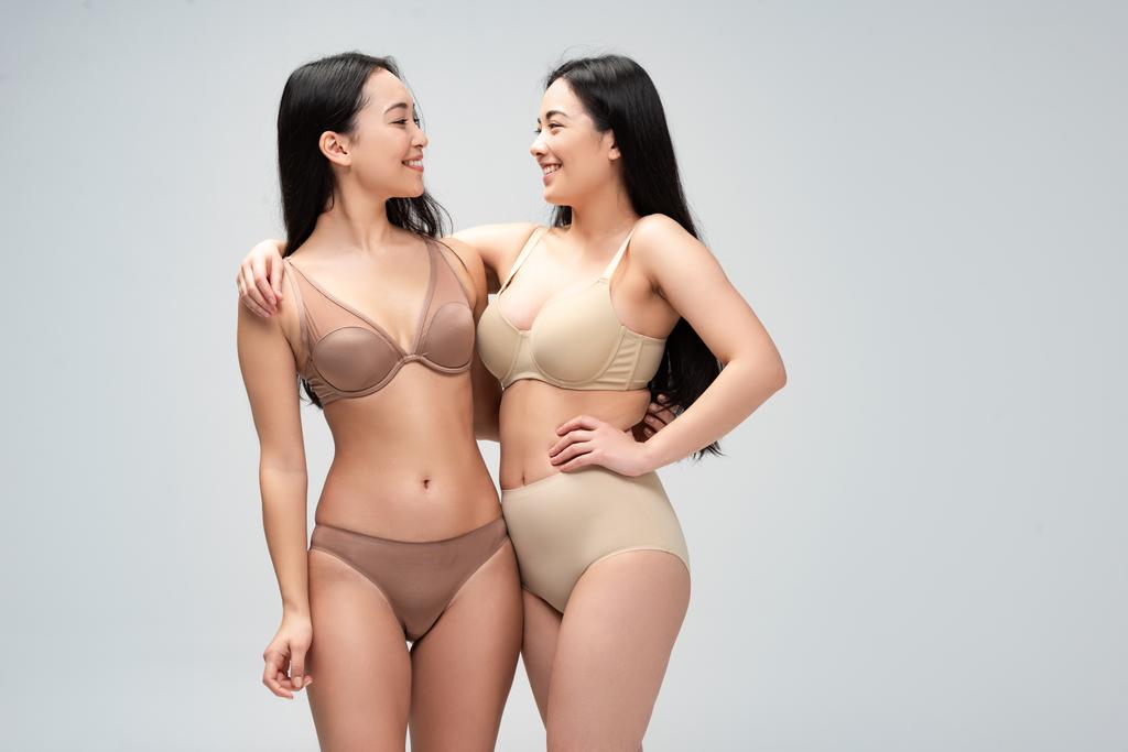 две весёлые многоэтнические женщины в нижнем белье обнимаются и смотрят друг на друга изолированно на сером, концепция позитивности тела
 - Фото, изображение
