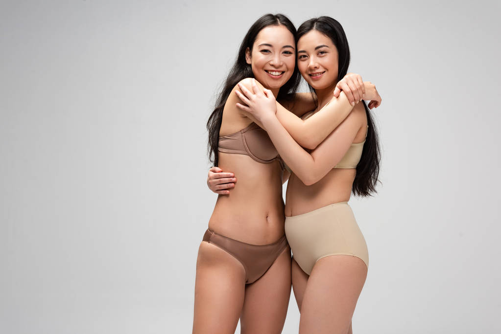 dos jóvenes y felices mujeres multiculturales abrazando y mirando a la cámara aislada en gris, concepto de positividad corporal
 - Foto, Imagen