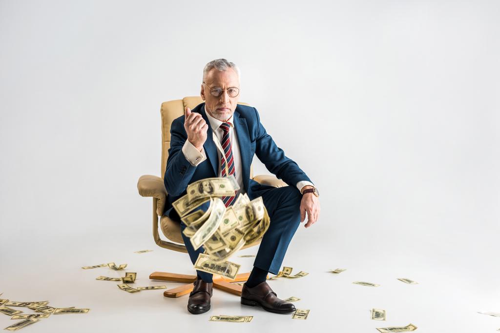 concentration sélective de l'homme d'affaires mûr sérieux assis dans un fauteuil et jetant des billets en dollars sur le gris
  - Photo, image
