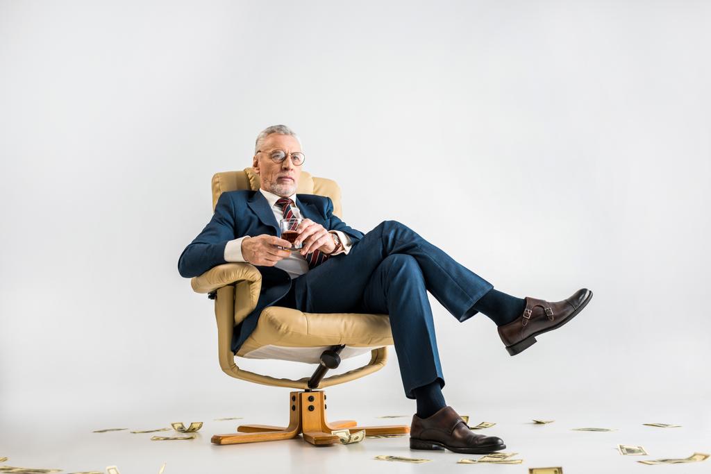 starší podnikatel v obleku sedí v křesle u dolarové bankovky a drží sklenici whisky Grey  - Fotografie, Obrázek