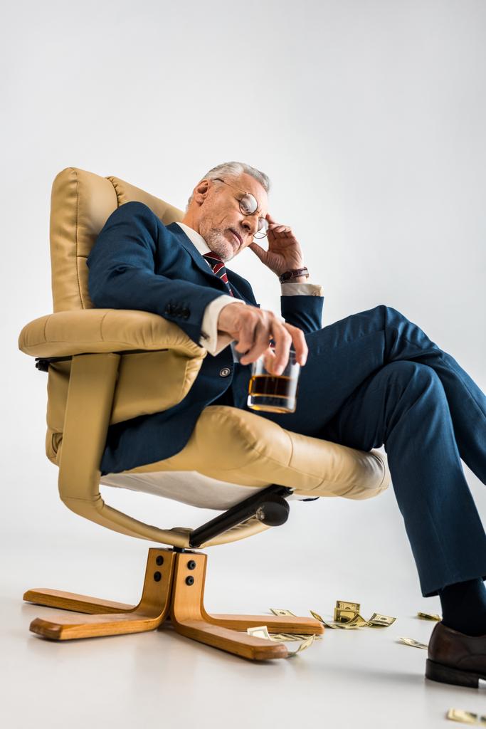 низький кут зору стомлюючий зрілий бізнесмен сидить у кріслі біля доларових купюр і тримає келих віскі на сірому
  - Фото, зображення