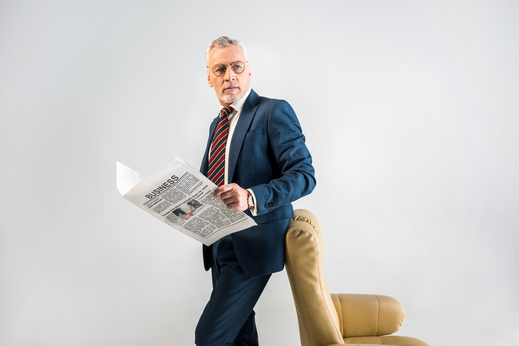 maduro hombre de negocios de pie cerca de sillón y la celebración de periódico de negocios aislado en gris
 - Foto, Imagen