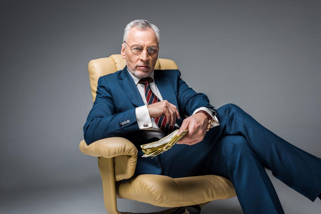 селективный фокус зрелого бизнесмена, сидящего в кресле и держащего долларовые банкноты на сером
  - Фото, изображение