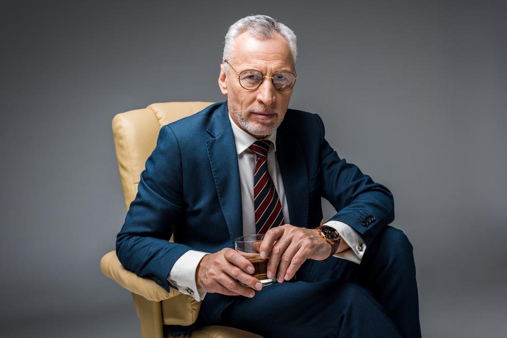 hombre de negocios maduro sentado en un sillón y sosteniendo un vaso de whisky en gris
 - Foto, Imagen