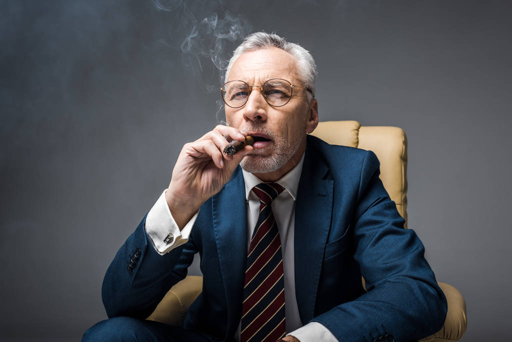 maduro hombre de negocios en traje y gafas de fumar cigarro en gris
 - Foto, Imagen