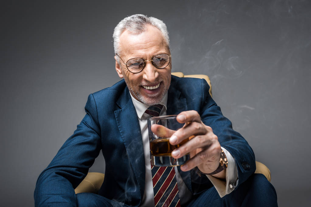 happy mature businessman in suit holding glass of whiskey while sitting in armchair on grey  - Photo, Image