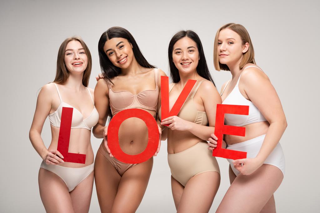 vier schöne multiethnische Mädchen in Unterwäsche, die das auf Papier geschnittene Wort "Liebe" isoliert auf grauem, körperpositivem Konzept halten  - Foto, Bild