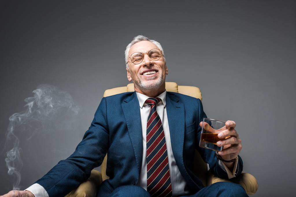 sorridente maduro empresário em terno segurando copo de uísque enquanto sentado em poltrona no cinza
  - Foto, Imagem