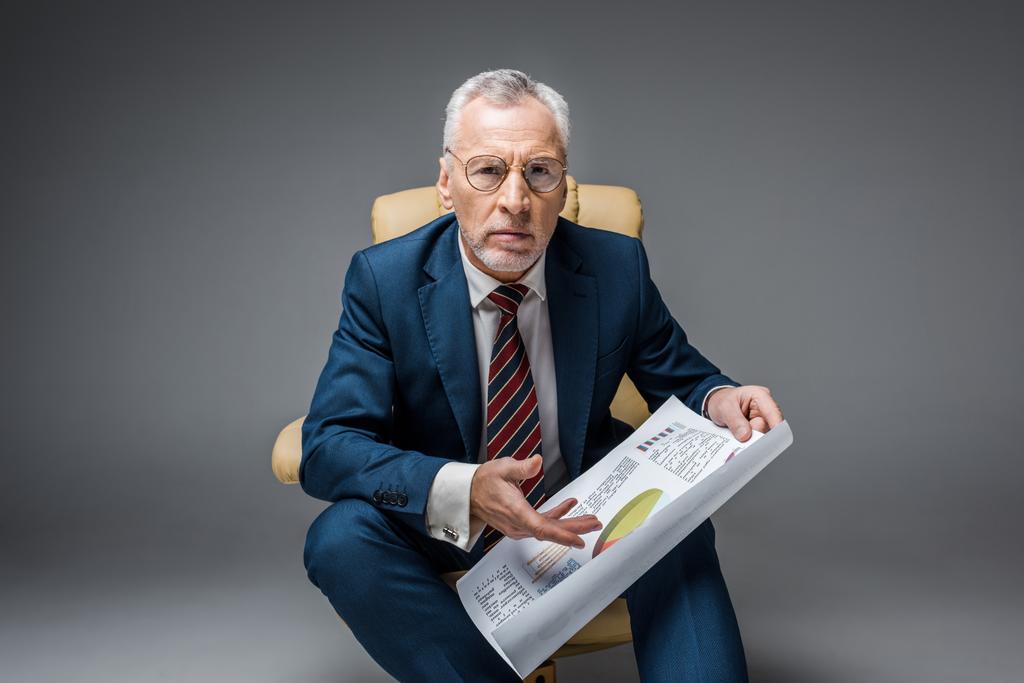 Reifer Geschäftsmann in Anzug und Brille mit Diagrammen und Grafiken im Sessel auf grau  - Foto, Bild