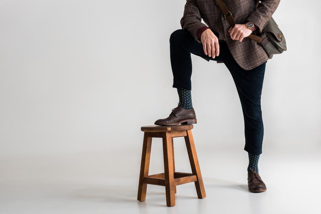 περικοπεί θέα κομψό ώριμο άνδρα που βάζει πόδι στην καρέκλα ενώ στέκεται στο γκρι  - Φωτογραφία, εικόνα