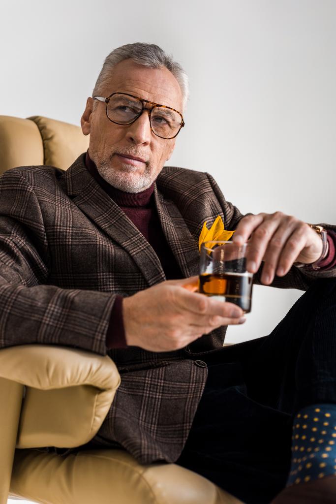 stílusos érett ember ül a karosszékben és a pohár whiskyt elszigetelt szürke gazdaság - Fotó, kép