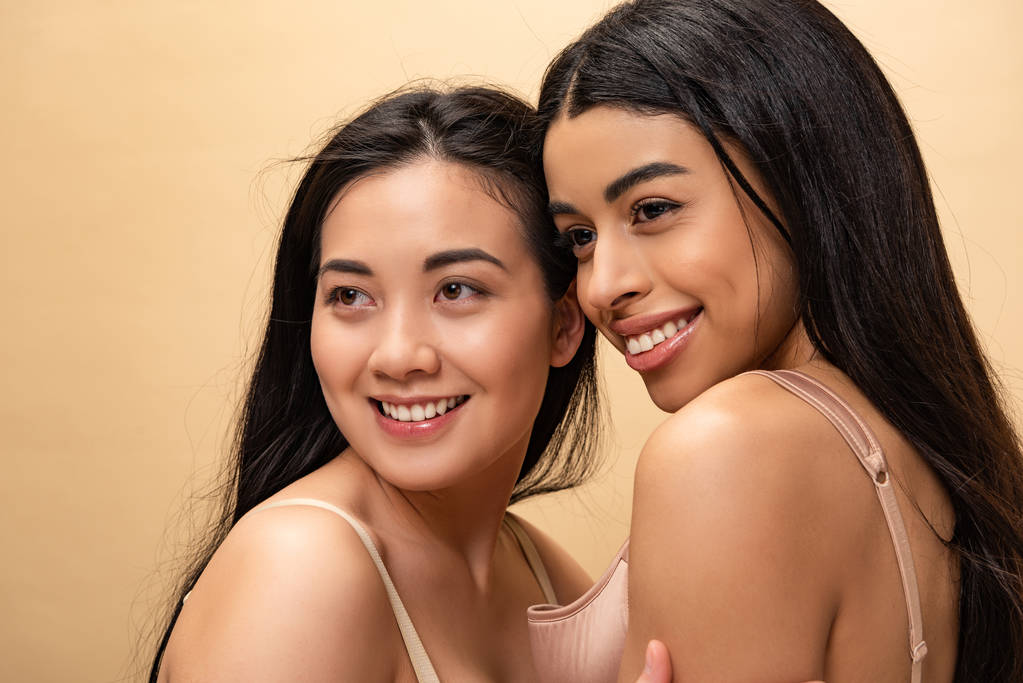 Kaksi kaunista monikulttuurista tyttöä hymyilee ja katsoo pois eristyksissä beige
 - Valokuva, kuva